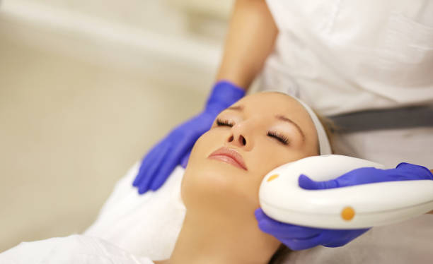 A practitioner in blue gloves using a smooth, handheld laser rejuvenation device on a woman's face during a skin treatment session.